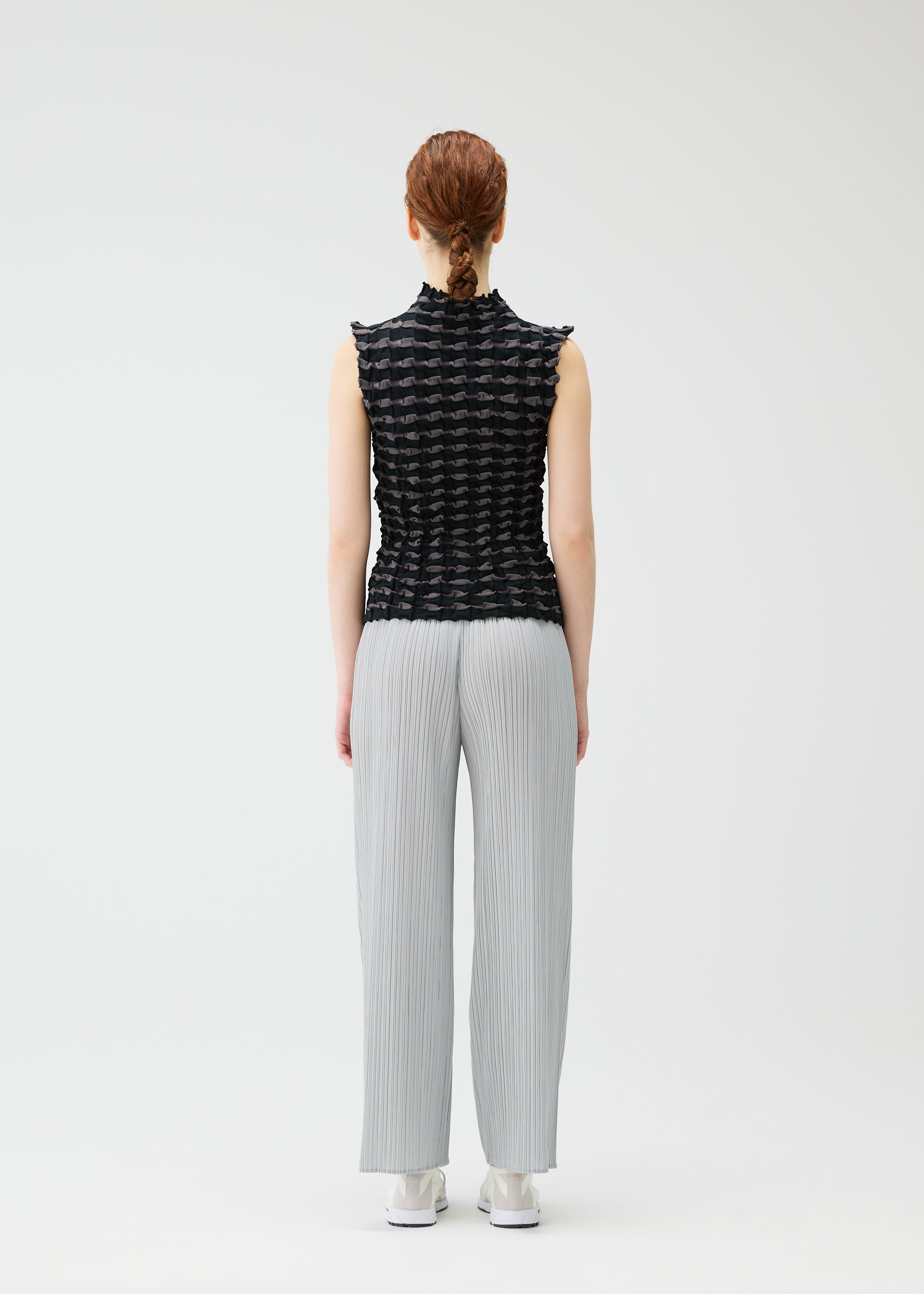 Zoomed view A person with reddish braided hair stands facing away, wearing the PLEATS PLEASE ISSEY MIYAKE TWIST KNIT top with light gray pleated pants and white shoes against a plain white background.