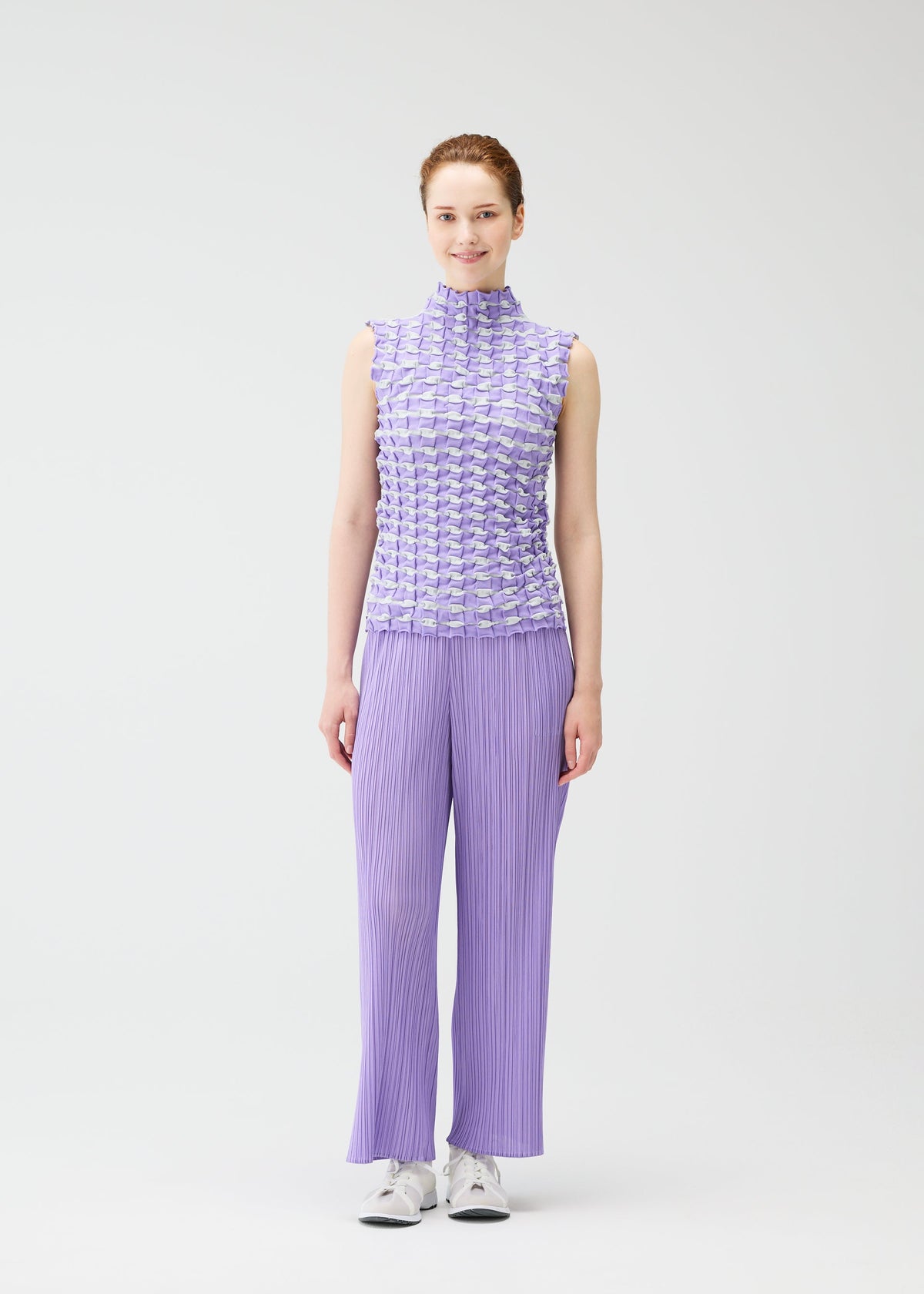 A woman faces forward, smiling, wearing the PLEATS PLEASE ISSEY MIYAKE TWIST KNIT sleeveless lavender top and wide-leg pants with white sneakers against a plain white background. view 8 of 8