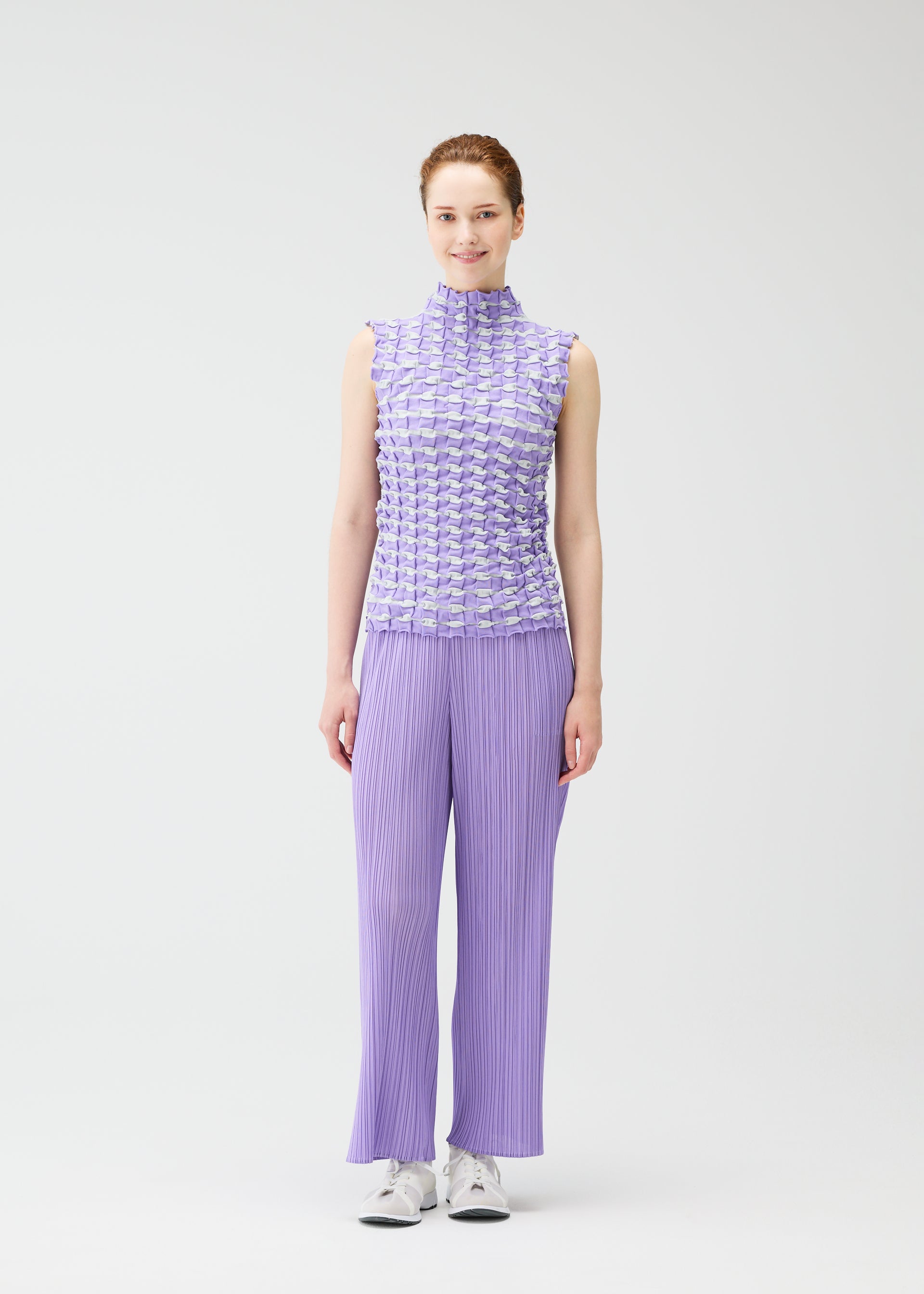 Zoomed view A woman faces forward, smiling, wearing the PLEATS PLEASE ISSEY MIYAKE TWIST KNIT sleeveless lavender top and wide-leg pants with white sneakers against a plain white background.