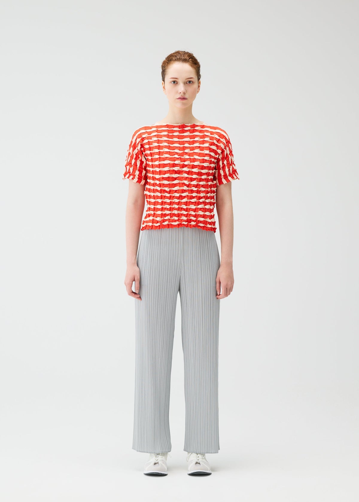 A person stands facing forward in a red and white TWIST KNIT top by PLEATS PLEASE ISSEY MIYAKE, light gray wide-leg pants, and white sneakers against a plain white background. view 5 of 7