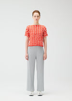 A person stands facing forward in a red and white TWIST KNIT top by PLEATS PLEASE ISSEY MIYAKE, light gray wide-leg pants, and white sneakers against a plain white background.