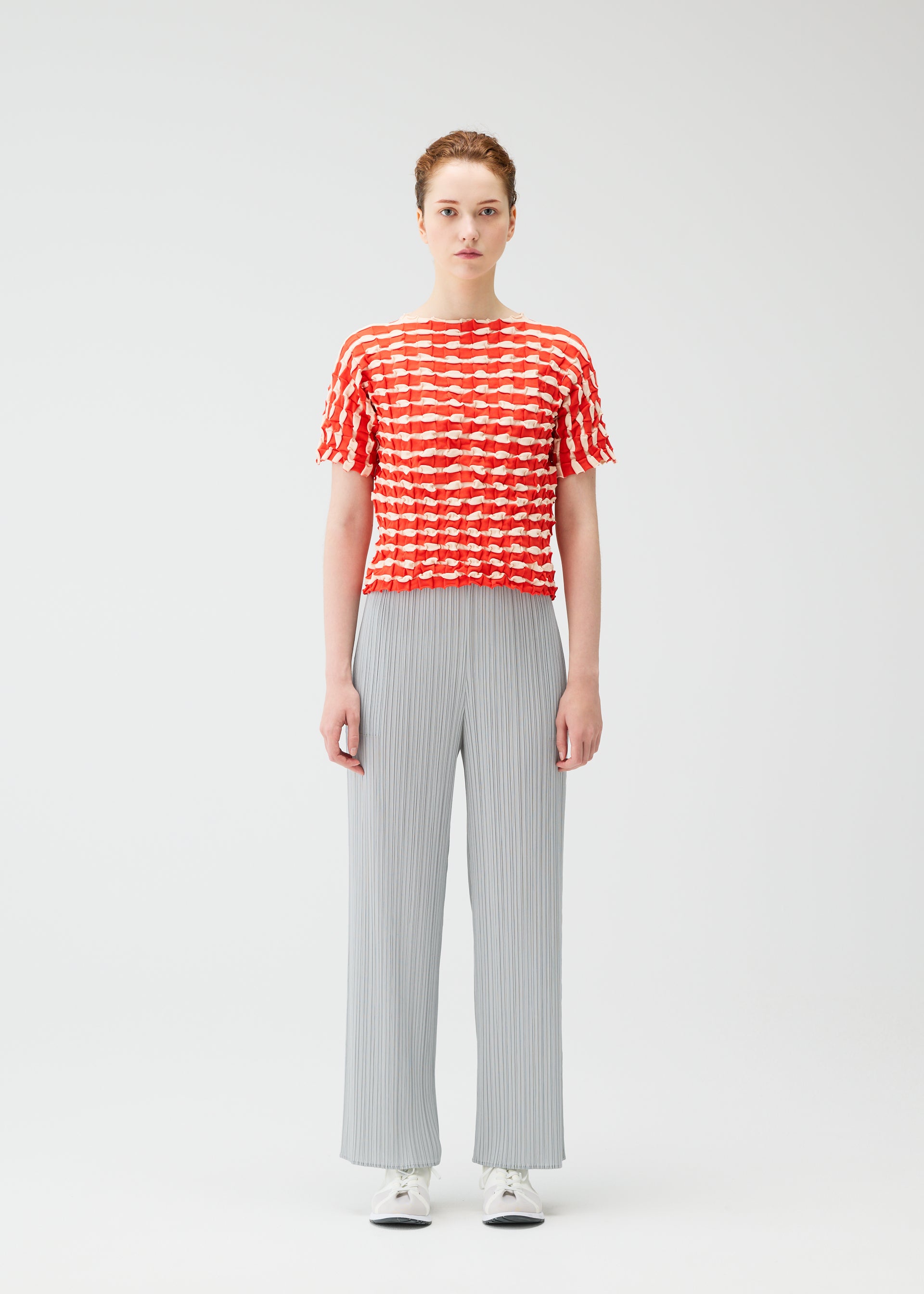 Zoomed view A person stands facing forward in a red and white TWIST KNIT top by PLEATS PLEASE ISSEY MIYAKE, light gray wide-leg pants, and white sneakers against a plain white background.
