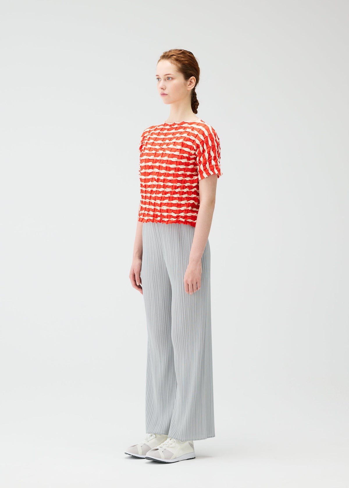 A woman with light skin and brown hair tied back stands sideways against a white background, wearing the PLEATS PLEASE ISSEY MIYAKE TWIST KNIT top, light gray pleated wide-leg pants, and white sneakers. view 6 of 7