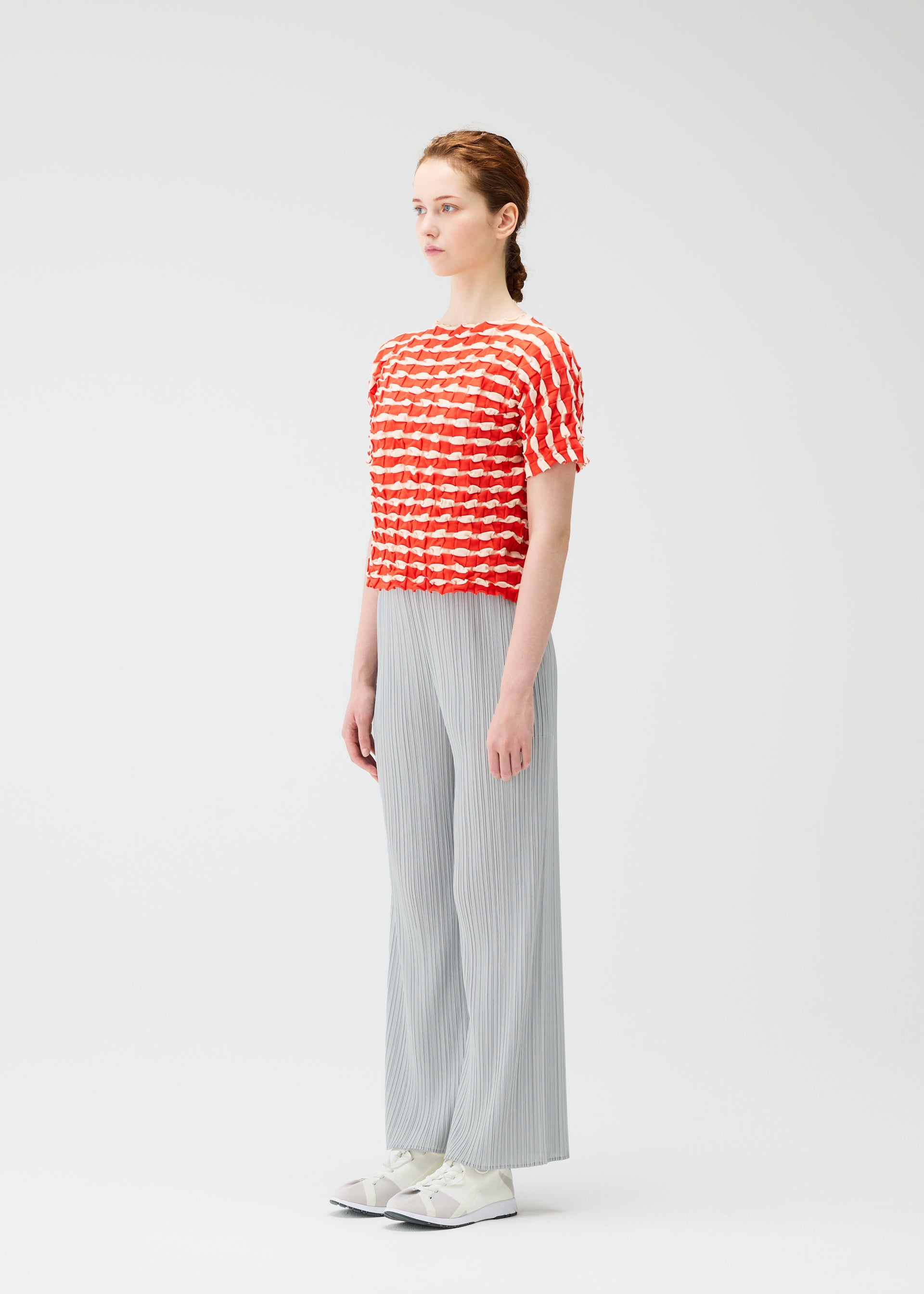 Zoomed view A woman with light skin and brown hair tied back stands sideways against a white background, wearing the PLEATS PLEASE ISSEY MIYAKE TWIST KNIT top, light gray pleated wide-leg pants, and white sneakers.