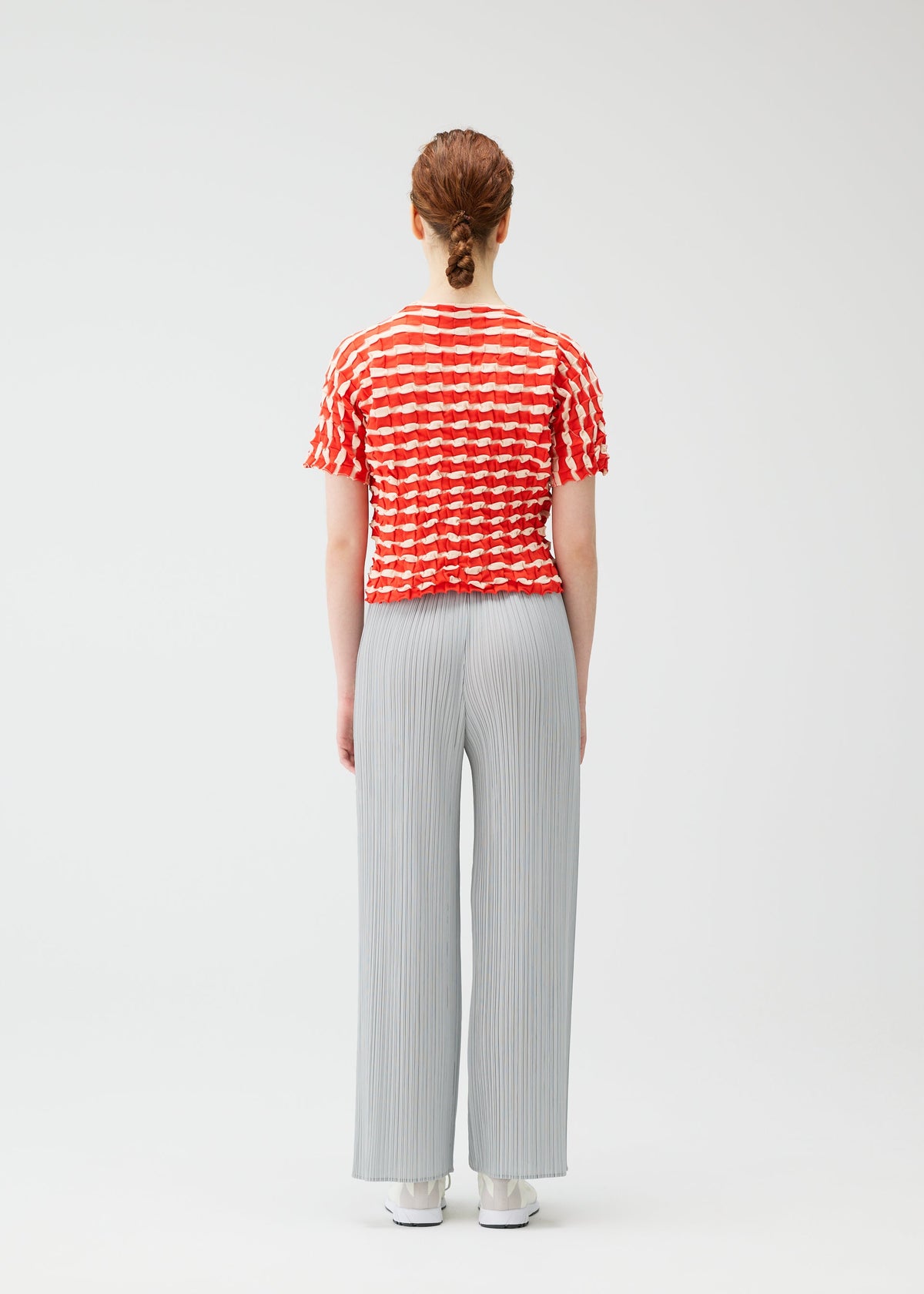 A person with auburn hair in a braid stands facing away, wearing the PLEATS PLEASE ISSEY MIYAKE TWIST KNIT top, light gray pleated wide-leg pants, and white shoes against a plain white background. view 7 of 7