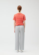 A person with auburn hair in a braid stands facing away, wearing the PLEATS PLEASE ISSEY MIYAKE TWIST KNIT top, light gray pleated wide-leg pants, and white shoes against a plain white background.