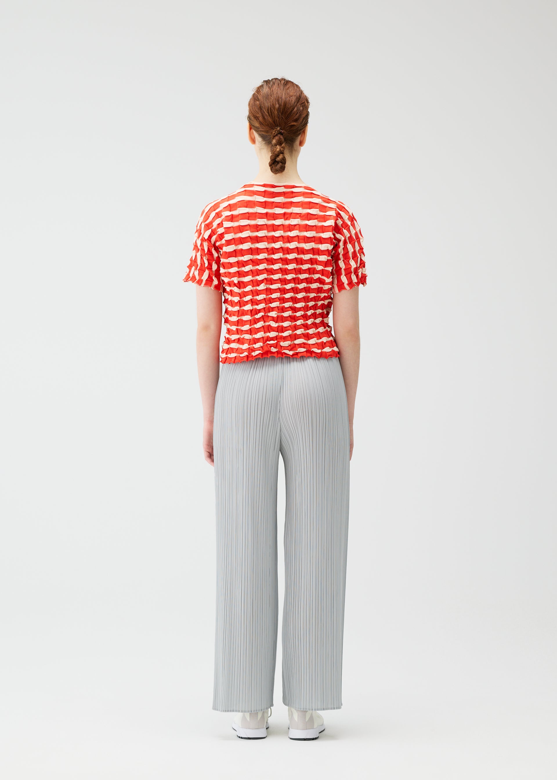 Zoomed view A person with auburn hair in a braid stands facing away, wearing the PLEATS PLEASE ISSEY MIYAKE TWIST KNIT top, light gray pleated wide-leg pants, and white shoes against a plain white background.
