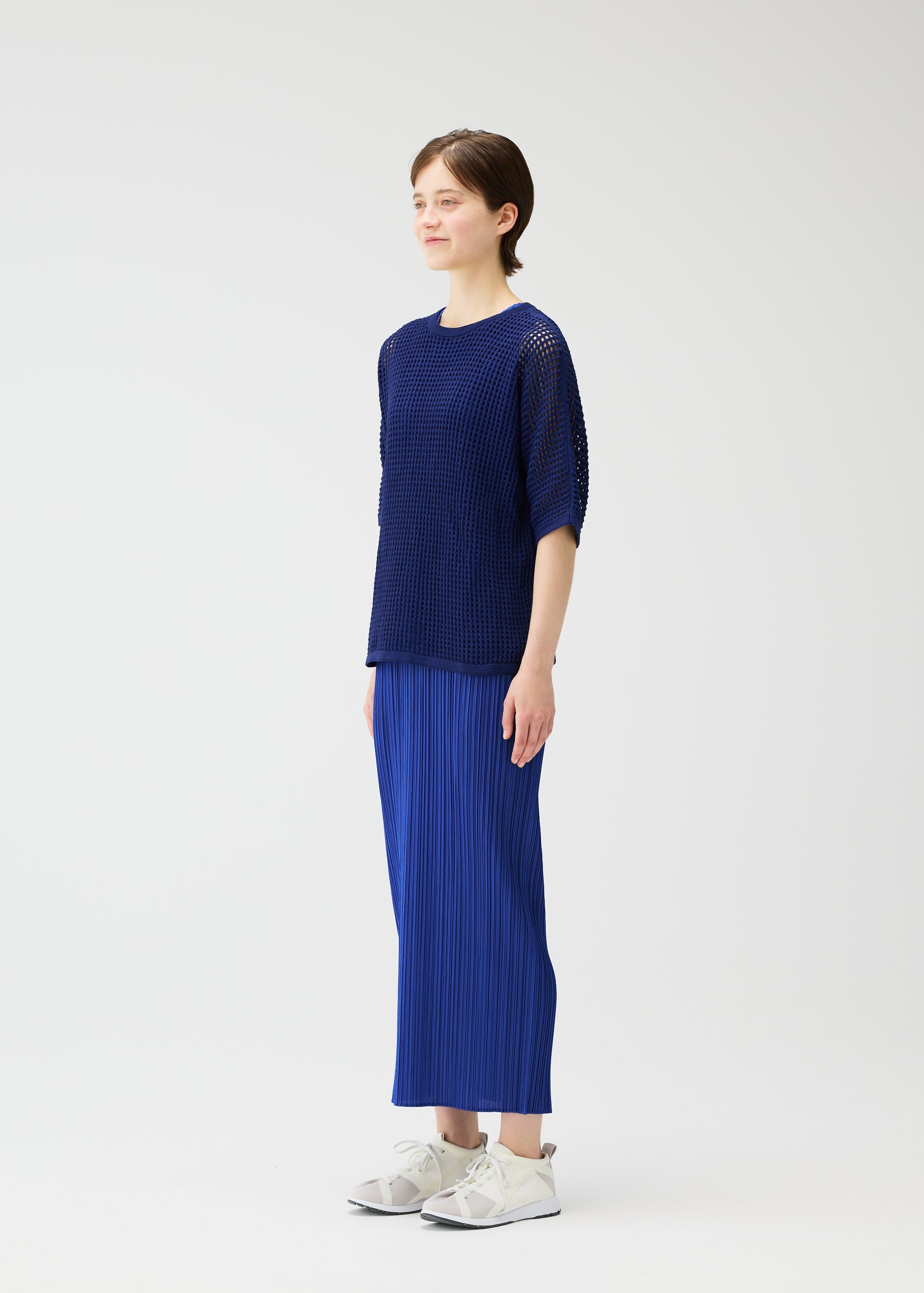 Zoomed view A person with short brown hair stands against a white background, wearing the PLEATS PLEASE ISSEY MIYAKE FISHNET KNIT top and matching pleated skirt in blue with white sneakers, looking slightly to the side and smiling softly.
