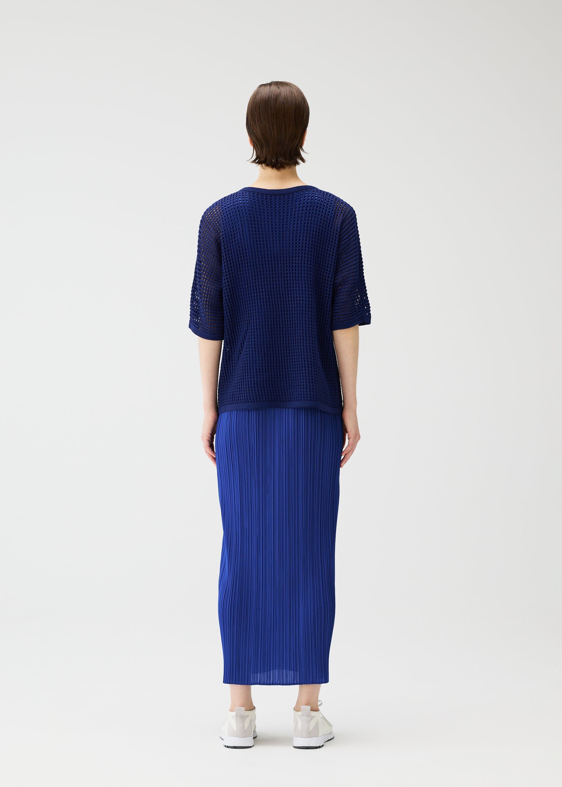 Zoomed view A person stands facing away, wearing the PLEATS PLEASE ISSEY MIYAKE FISHNET KNIT top and a matching pleated skirt in dark blue with white sneakers, set against a plain light background.