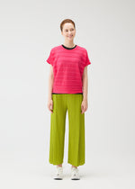 A smiling person poses against a plain white background in the STEP KNIT top and loose lime green pleated pants by PLEATS PLEASE ISSEY MIYAKE, paired with white sneakers—an ideal summer look.