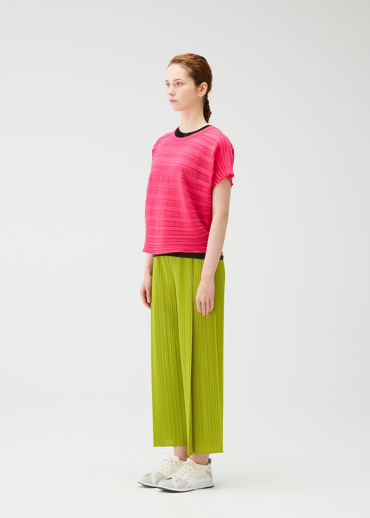 A woman stands in profile against a white background, wearing the STEP KNIT top and lime green pleated pants from PLEATS PLEASE ISSEY MIYAKE—a striking summer outfit paired with white sneakers. view 6 of 7