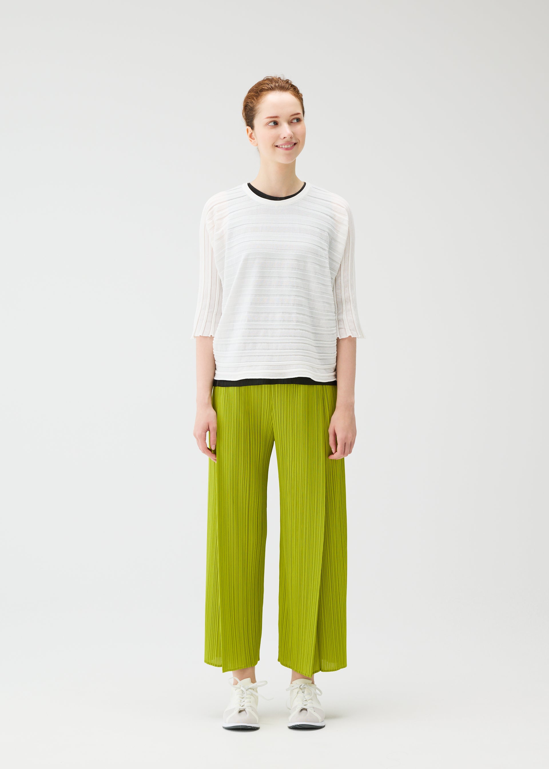 Zoomed view Against a plain white background, a person smiles while wearing the STEP KNIT top and wide-legged bright green pleated pants by PLEATS PLEASE ISSEY MIYAKE with white sneakers—an effortlessly fresh summer look.