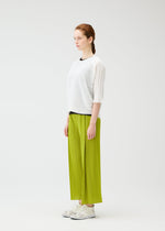 A woman with braided hair stands in profile against a white background, wearing the PLEATS PLEASE ISSEY MIYAKE STEP KNIT white top and lime green pleated wide-leg pants—an effortless summer look completed with white sneakers.