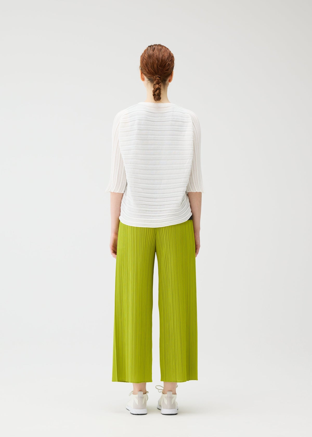 A person with braided red hair faces away, wearing the PLEATS PLEASE ISSEY MIYAKE STEP KNIT top, loose bright green pleated pants, and white sneakers against a plain white background. view 7 of 7