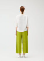 A person with braided red hair faces away, wearing the PLEATS PLEASE ISSEY MIYAKE STEP KNIT top, loose bright green pleated pants, and white sneakers against a plain white background.