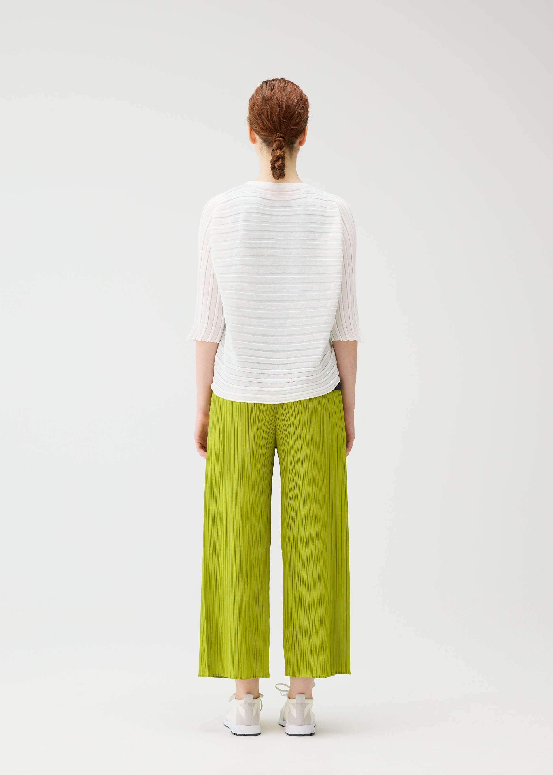 Zoomed view A person with braided red hair faces away, wearing the PLEATS PLEASE ISSEY MIYAKE STEP KNIT top, loose bright green pleated pants, and white sneakers against a plain white background.