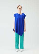 A woman with light skin and auburn hair tied back stands against a white background, wearing the PLEATS PLEASE ISSEY MIYAKE A-POC SEA GLASS tunic in bright blue, teal pleated pants, and white sneakers, smiling slightly.