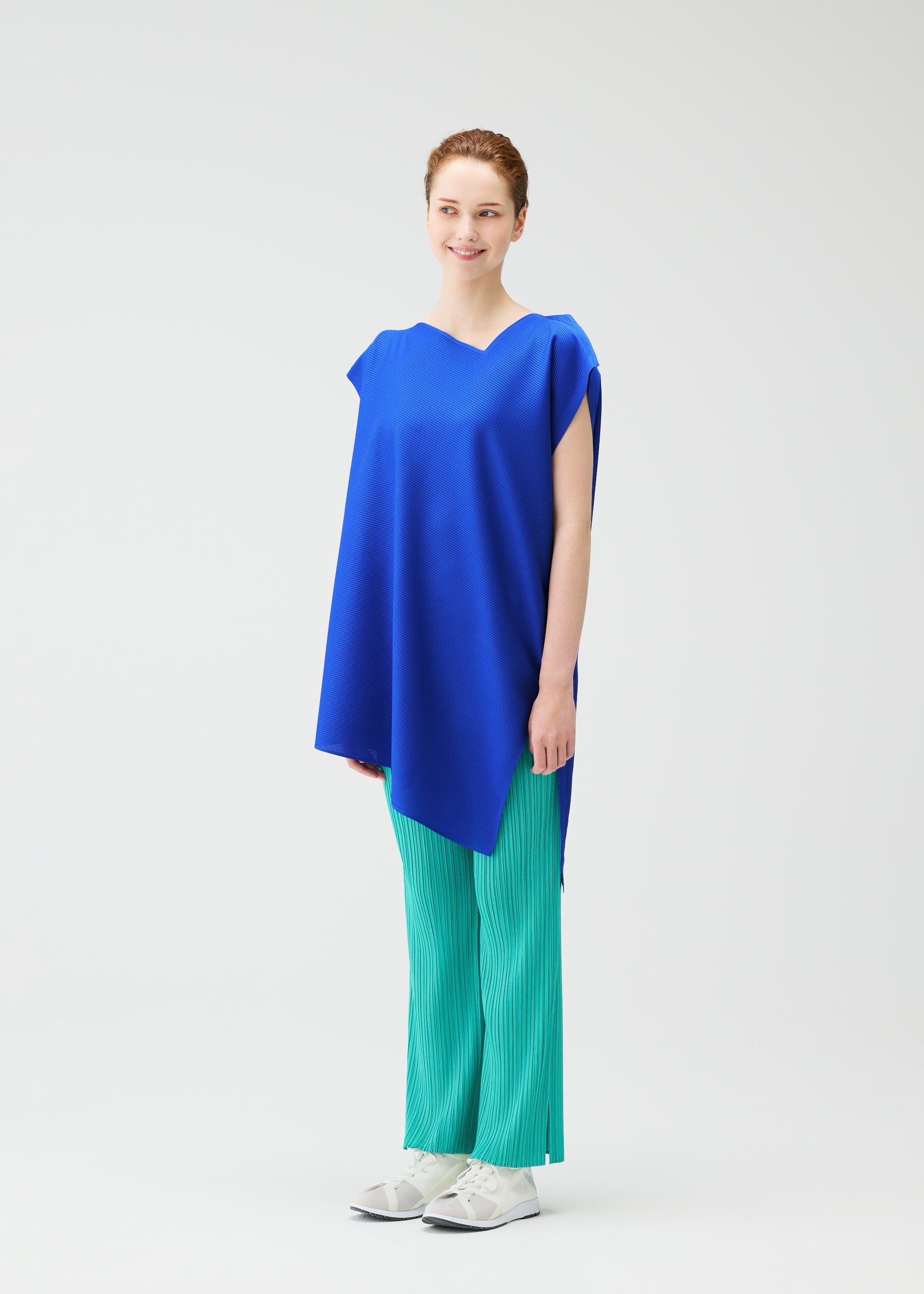 Zoomed view A woman smiles, wearing the PLEATS PLEASE ISSEY MIYAKE A-POC SEA GLASS tunic in blue with an asymmetrical hem, paired with green pleated pants and white sneakers, set against a plain white background.