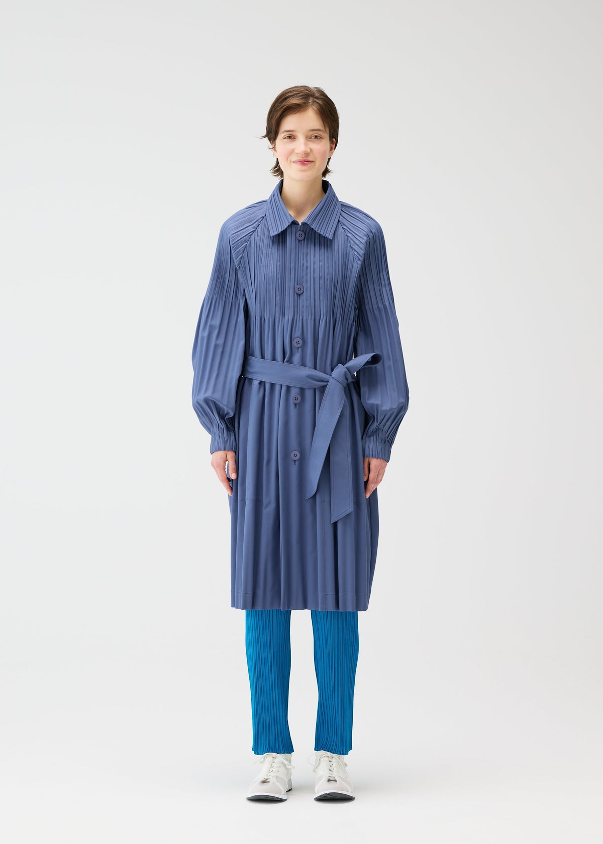 A person faces forward in a PLEATS PLEASE ISSEY MIYAKE SMOOTH COAT, a wind-resistant blue trench with Keshi pleats and matching belt, bright blue pleated pants, and white shoes, against a plain white background. view 6 of 9