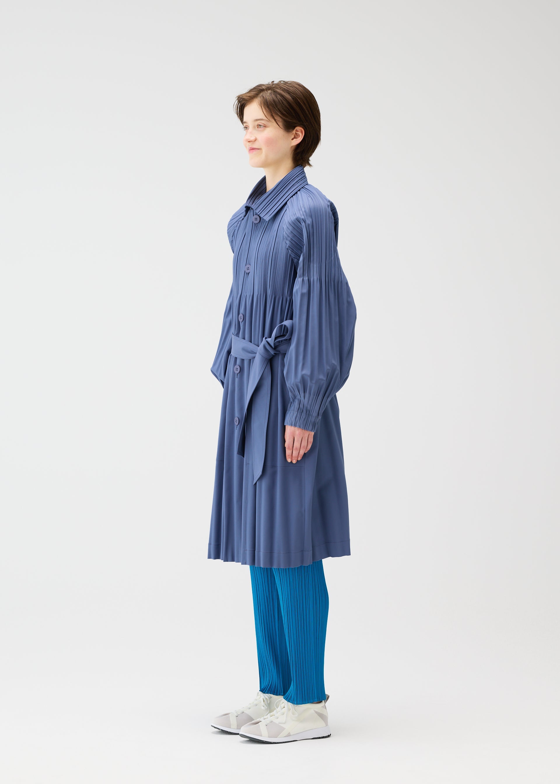 Zoomed view A person stands sideways wearing a long-sleeved, belted SMOOTH COAT by PLEATS PLEASE ISSEY MIYAKE, blue pleated pants, and white shoes against a plain white background.