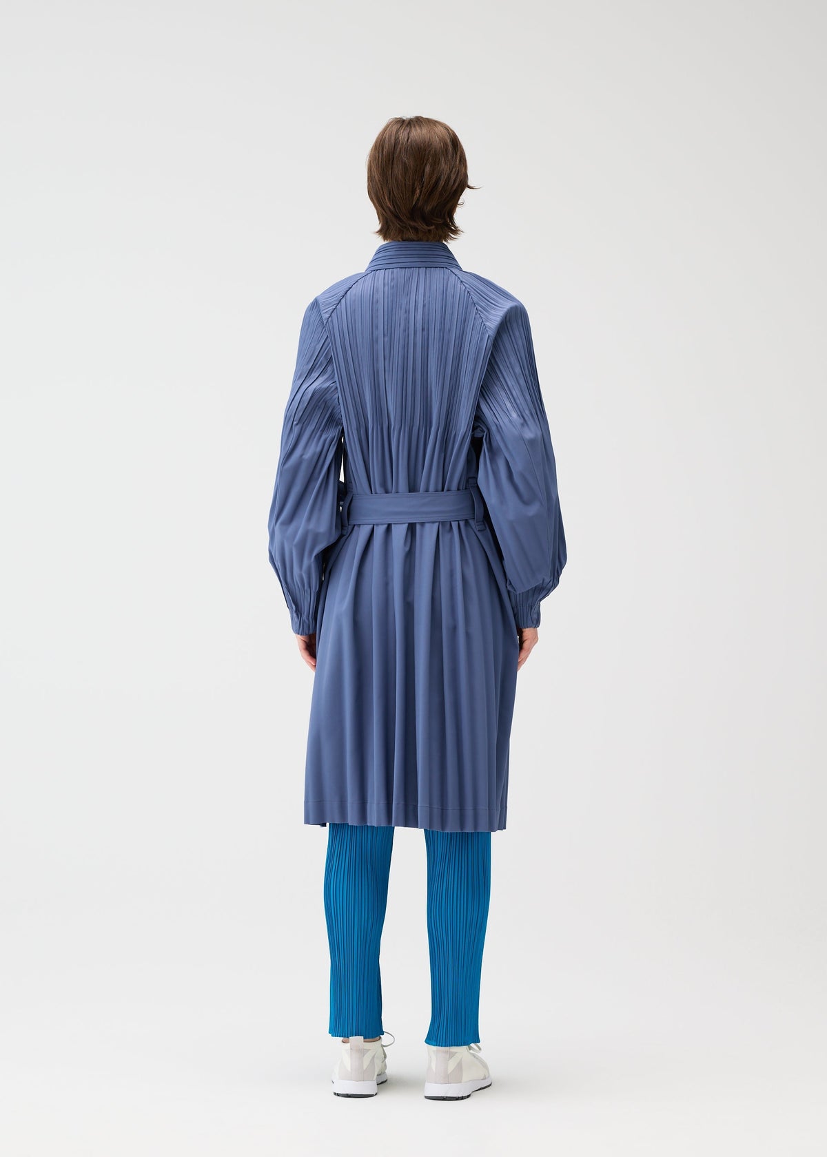 A person faces away, wearing the SMOOTH COAT by PLEATS PLEASE ISSEY MIYAKE in blue with a belt, paired with bright blue pleated tights and white shoes against a plain light background. view 8 of 9