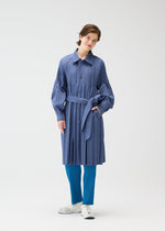 A person wearing the blue SMOOTH COAT by PLEATS PLEASE ISSEY MIYAKE over bright blue Keshi pleats pants and white sneakers stands against a plain white background, smiling slightly.