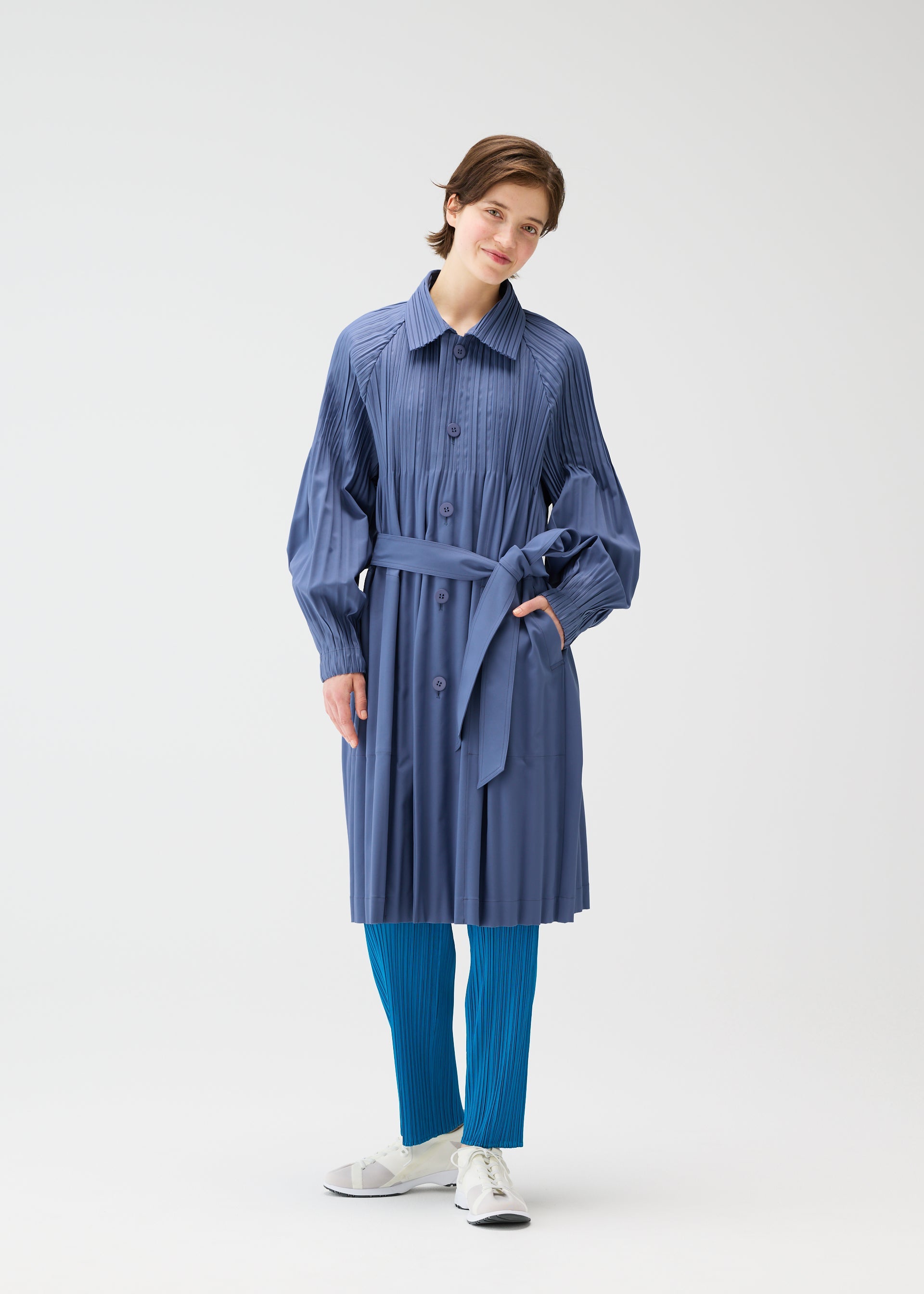 Zoomed view A person wearing the blue SMOOTH COAT by PLEATS PLEASE ISSEY MIYAKE over bright blue Keshi pleats pants and white sneakers stands against a plain white background, smiling slightly.