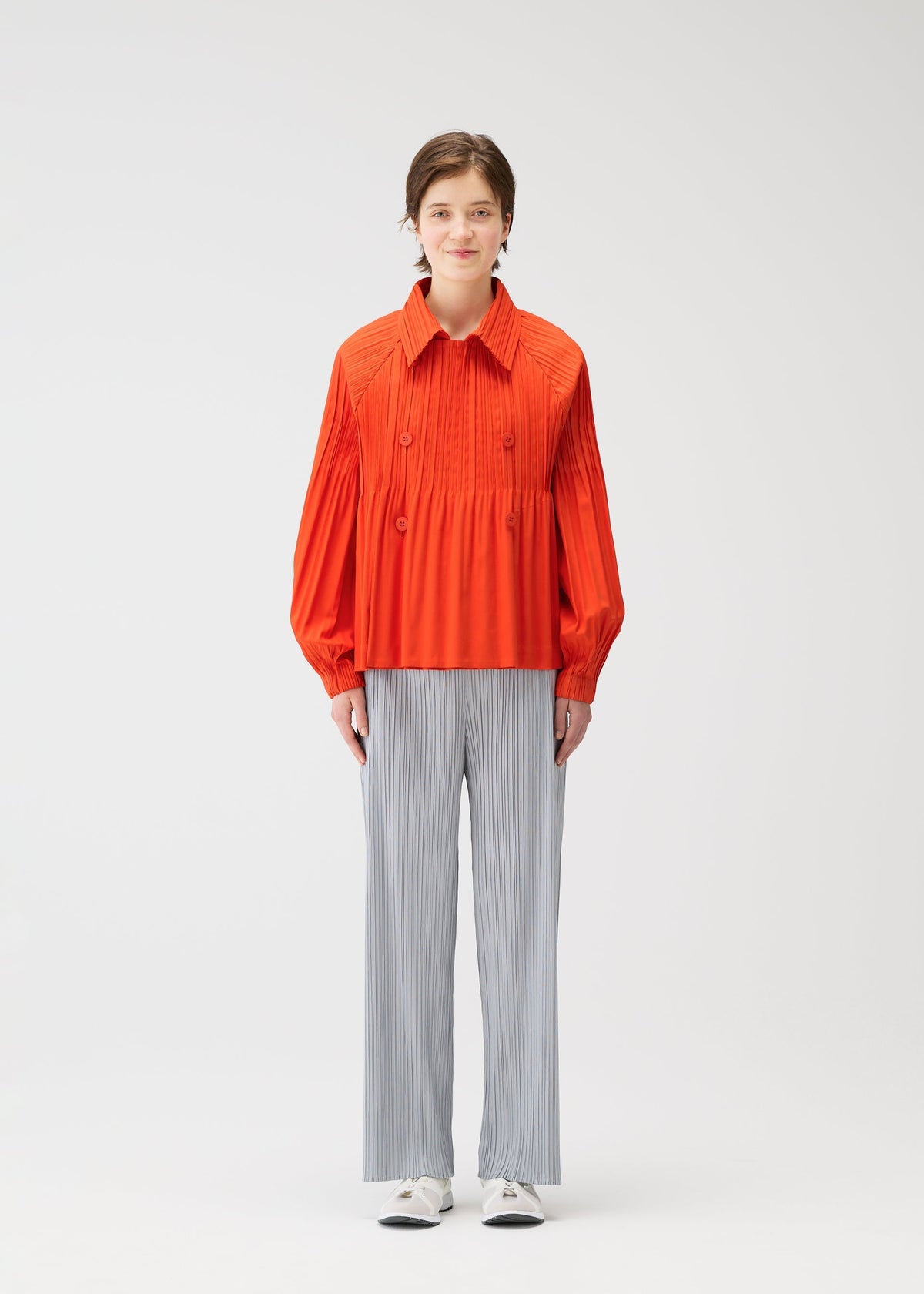 A person stands against a plain white background wearing the bright orange SMOOTH COAT by PLEATS PLEASE ISSEY MIYAKE, paired with loose, light gray pinstriped pants and white shoes. view 6 of 10