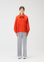 A person stands against a plain white background wearing the bright orange SMOOTH COAT by PLEATS PLEASE ISSEY MIYAKE, paired with loose, light gray pinstriped pants and white shoes.