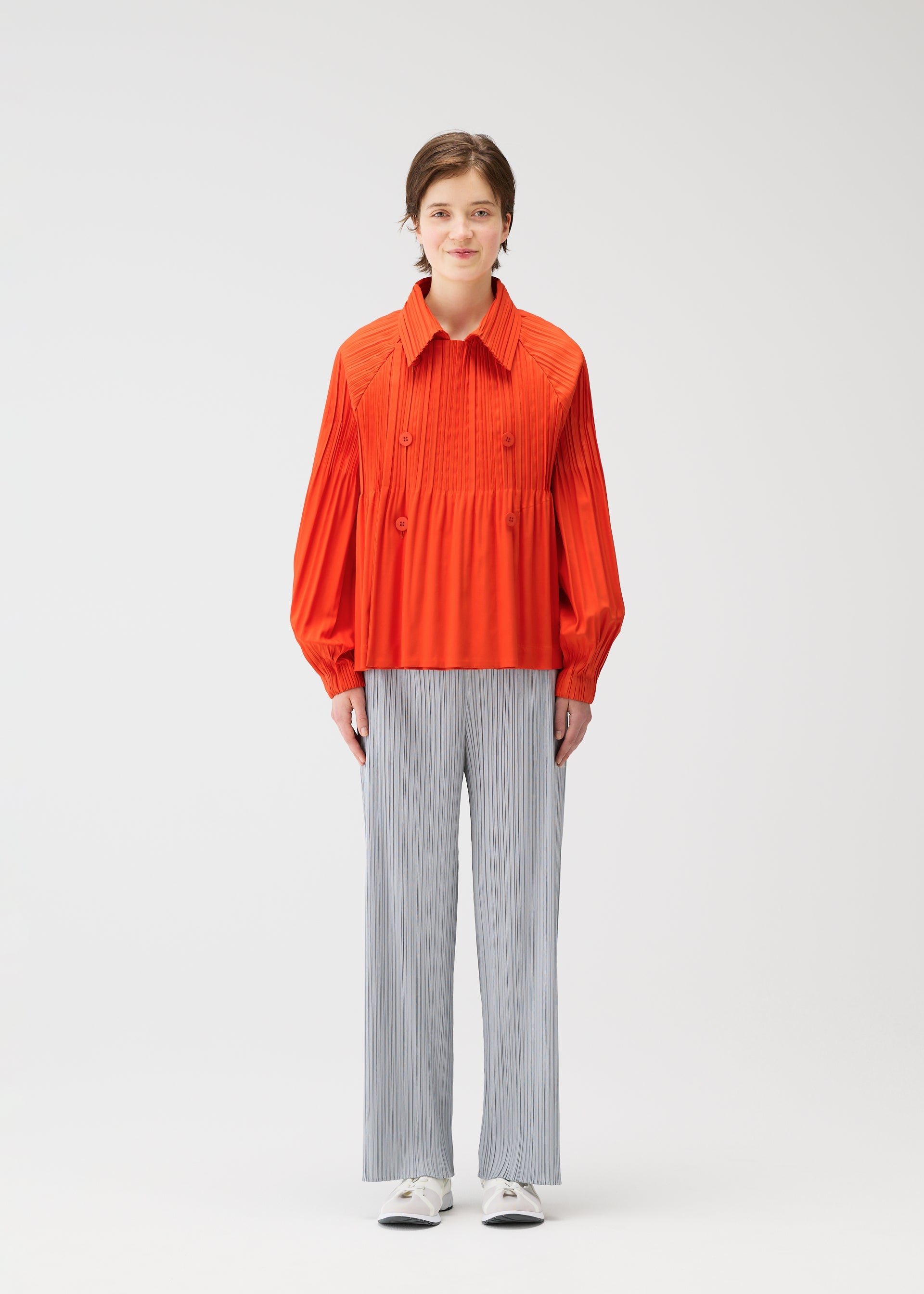 Zoomed view A person stands against a plain white background wearing the bright orange SMOOTH COAT by PLEATS PLEASE ISSEY MIYAKE, paired with loose, light gray pinstriped pants and white shoes.