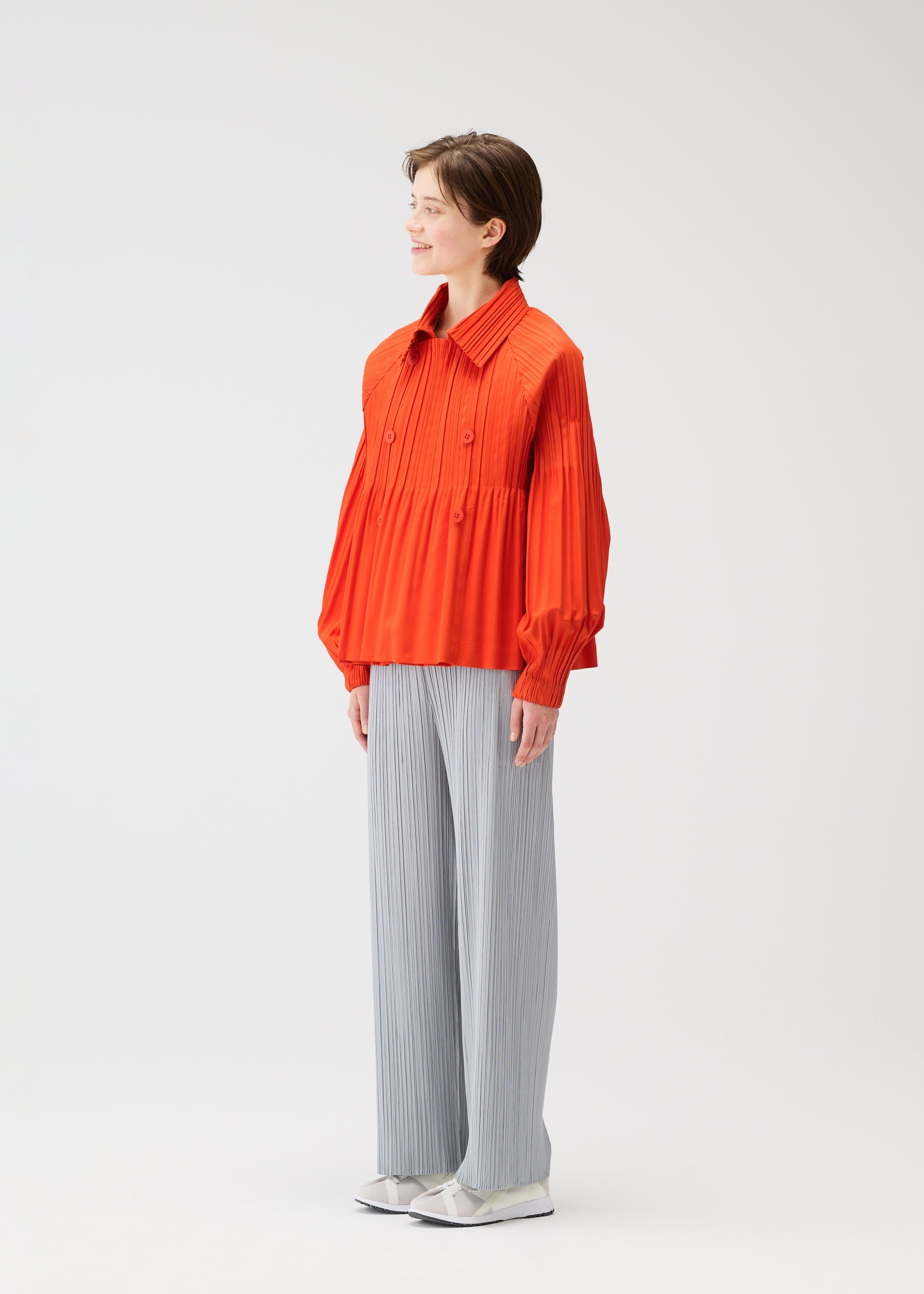 Zoomed view A person stands smiling in a SMOOTH COAT by PLEATS PLEASE ISSEY MIYAKE, paired with light gray wide-leg pants and white shoes, against a plain light background.
