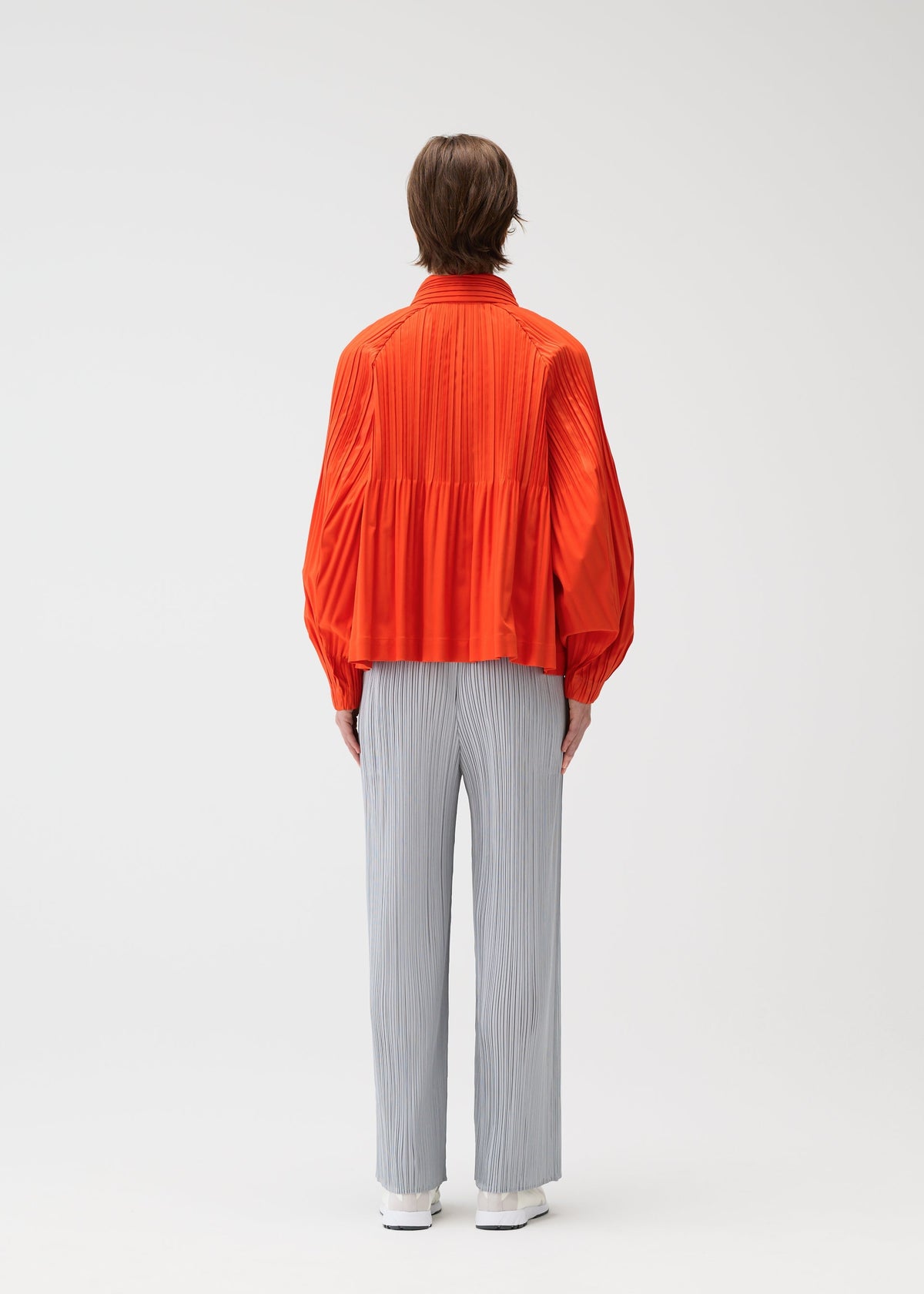 A person stands facing away in a bright orange SMOOTH COAT by PLEATS PLEASE ISSEY MIYAKE with Keshi pleats and voluminous sleeves, paired with light gray straight-leg pants against a plain white background. view 8 of 10