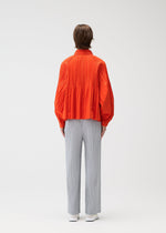 A person stands facing away in a bright orange SMOOTH COAT by PLEATS PLEASE ISSEY MIYAKE with Keshi pleats and voluminous sleeves, paired with light gray straight-leg pants against a plain white background.