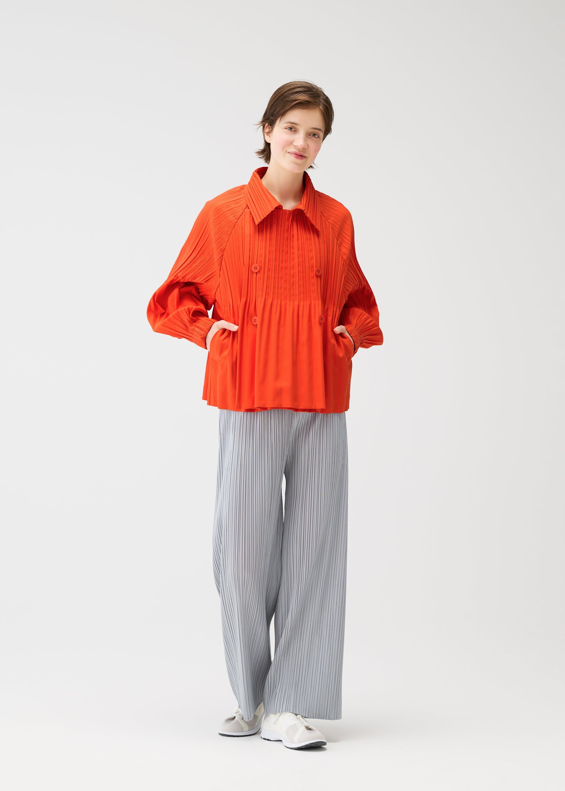 Zoomed view A person stands against a plain white background wearing the SMOOTH COAT by PLEATS PLEASE ISSEY MIYAKE, paired with light gray pinstriped pants and white shoes, hands in pockets, smiling softly.