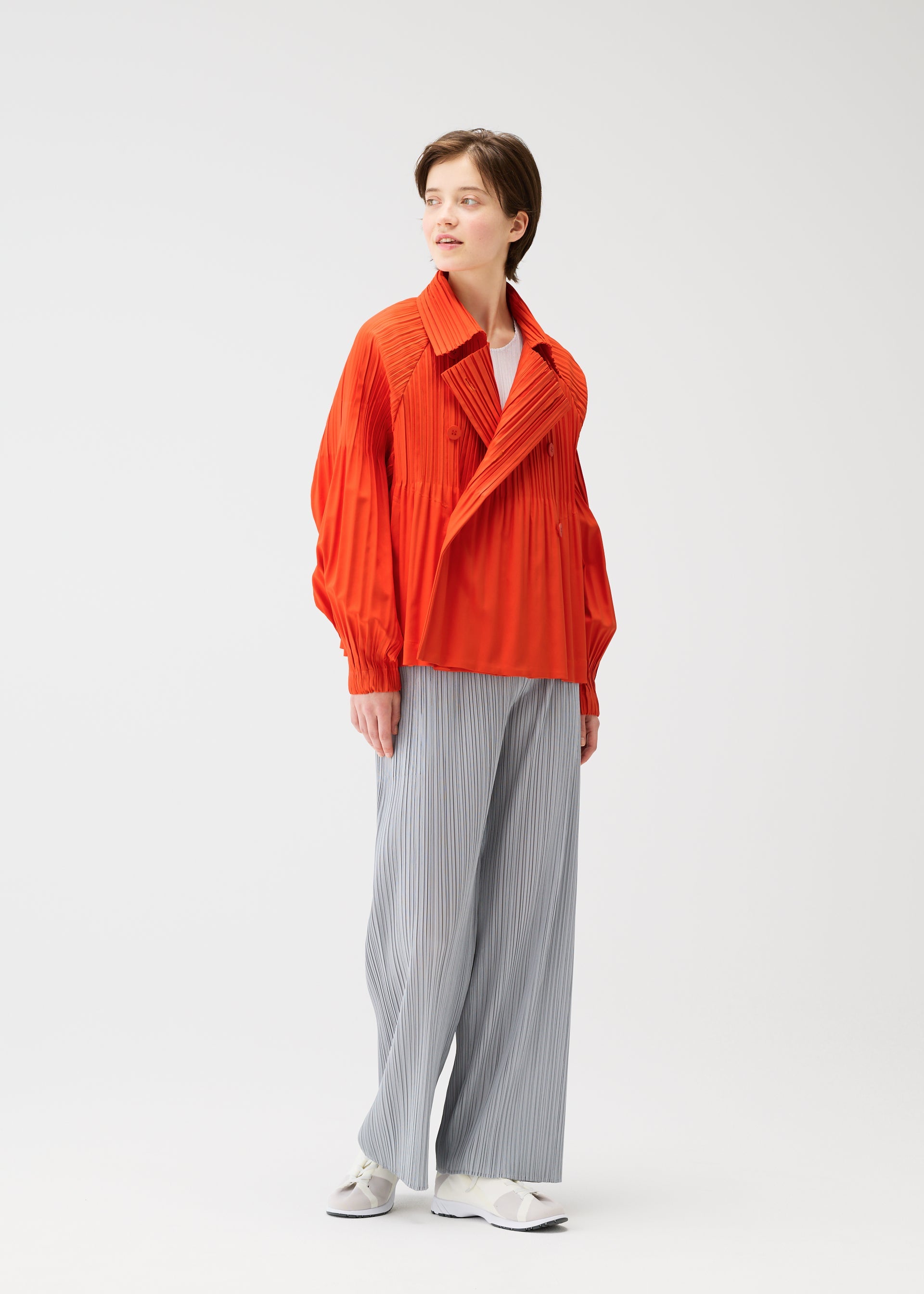 Zoomed view A person stands against a plain white background wearing a bright orange SMOOTH COAT from PLEATS PLEASE ISSEY MIYAKE, light gray wide-leg striped pants, and white sneakers.
