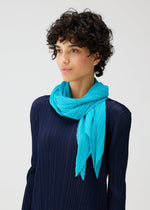 A person with short curly hair wears a navy pleated top and the MONTHLY SCARF JANUARY by PLEATS PLEASE ISSEY MIYAKE, standing against a plain light background like a minimalist folding screen.