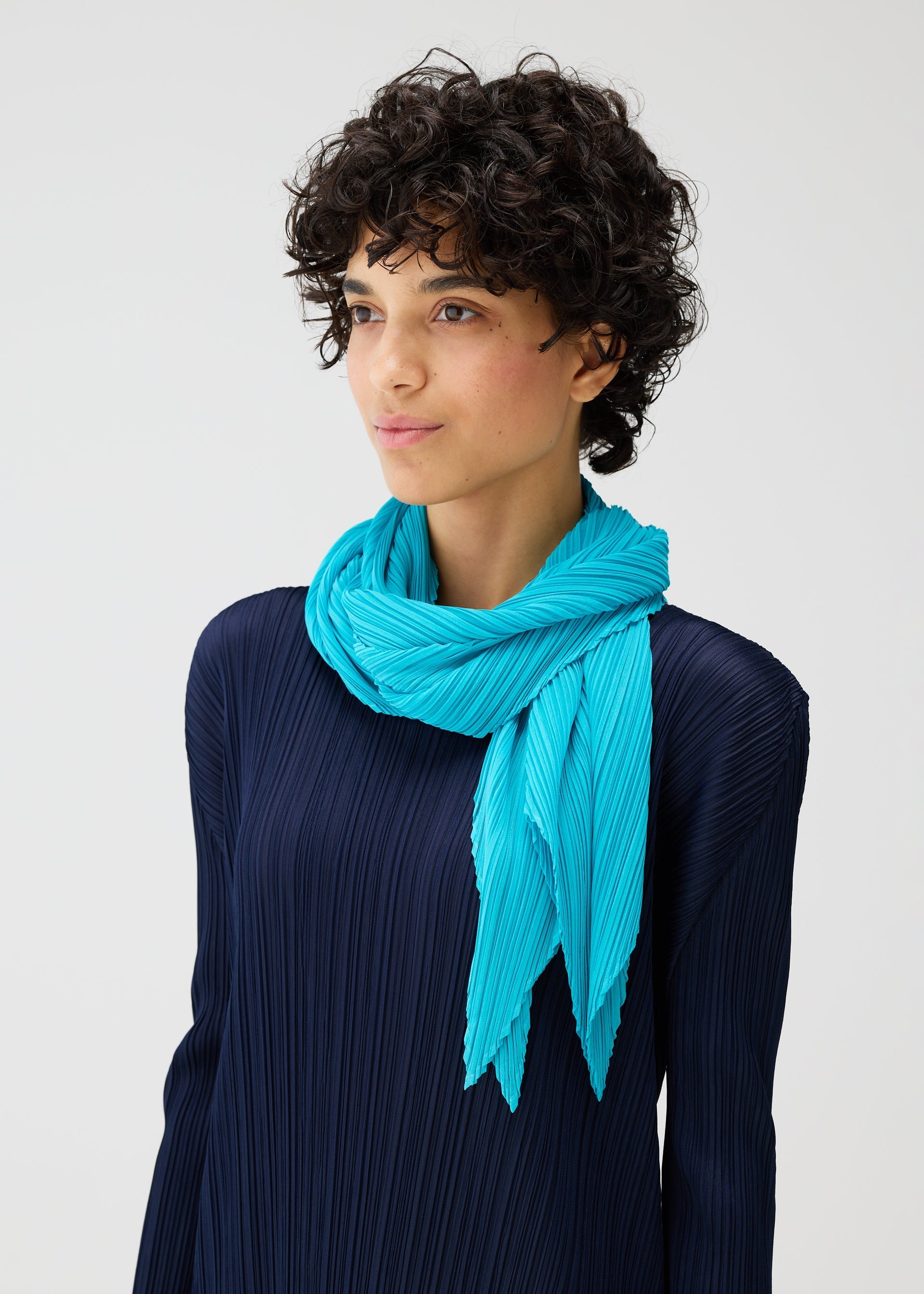 Zoomed view A person with short curly hair wears a navy pleated top and the MONTHLY SCARF JANUARY by PLEATS PLEASE ISSEY MIYAKE, standing against a plain light background like a minimalist folding screen.
