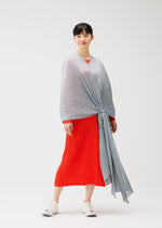 A person stands against a plain background in the bright red MADAME-T FEBRUARY dress by PLEATS PLEASE ISSEY MIYAKE, featuring bold pleats and a large stole tied in front. They have short black hair and wear white sneakers for a vivid, stylish look.