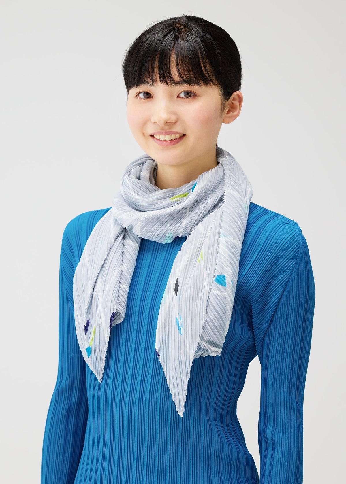A young person with straight black hair and bangs smiles at the camera, wearing a bright blue ribbed top and the PLEATS PLEASE ISSEY MIYAKE CROSSING DOTS SCARF against a plain light background. view 5 of 5