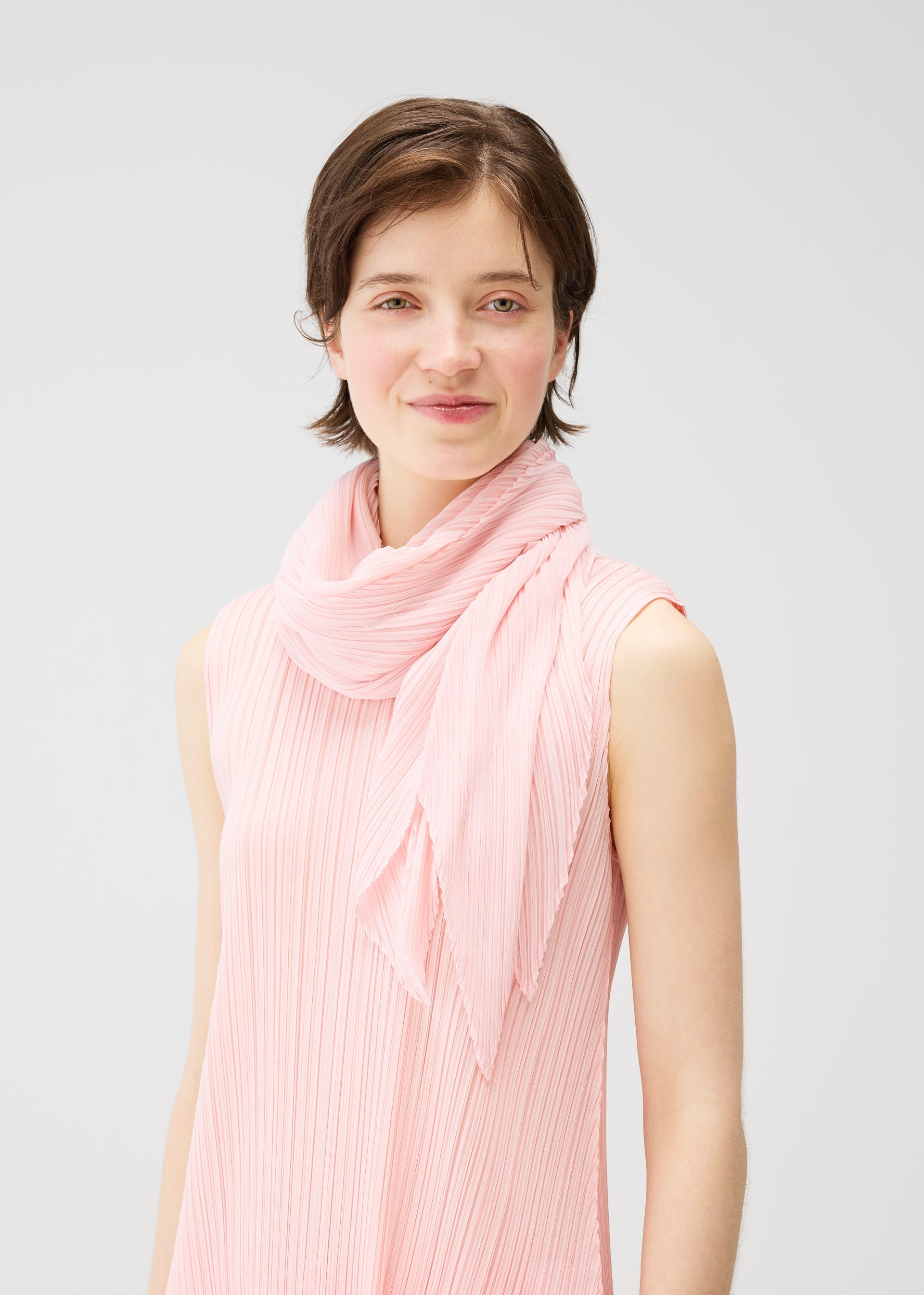 Zoomed view A person with short brown hair smiles softly, wearing the PLEATS PLEASE ISSEY MIYAKE MONTHLY SCARF MARCH and a matching sleeveless, light pink pleated top, posed before a folding screen against a plain light background.
