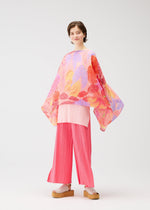 A person with short brown hair and platform sandals stands before a white background, wearing the FOGGY FOREST MADAME-T by PLEATS PLEASE ISSEY MIYAKE: wide-legged pink pleated pants, a light pink top, and a loose colorful printed stole.