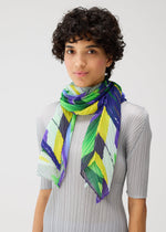 A person with short curly hair wears the PLAYGROUND SCARF by PLEATS PLEASE ISSEY MIYAKE—a pleated geometric print in green, yellow, blue, and white chevrons—over a light gray ribbed top against a plain background.