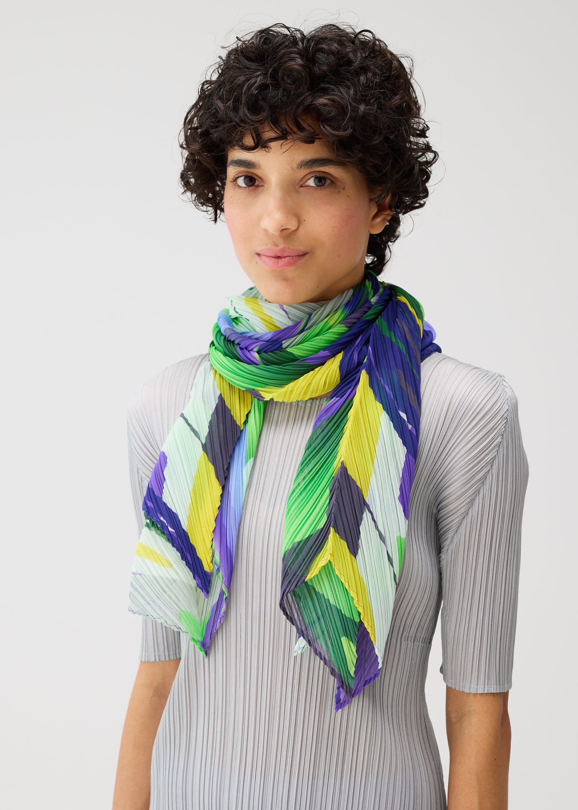 Zoomed view A person with short curly hair wears the PLAYGROUND SCARF by PLEATS PLEASE ISSEY MIYAKE—a pleated geometric print in green, yellow, blue, and white chevrons—over a light gray ribbed top against a plain background.