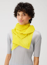 Person with short curly dark hair wearing a light gray ribbed top and the ZIGZAG SCARF by PLEATS PLEASE ISSEY MIYAKE, a bright yellow pleated scarf with a subtle zigzag pattern, standing against a plain white background.