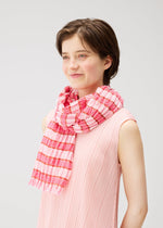 A person with short brown hair smiles while wearing a sleeveless, light pink top and the COTTON CHECK SCARF by PLEATS PLEASE ISSEY MIYAKE. The background is plain and light-colored.