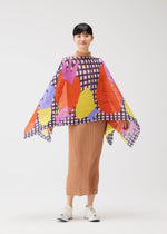 A woman smiles in a pleated tan skirt and white sneakers, wearing the PLEATS PLEASE ISSEY MIYAKE MARKET MADAME-T—a colorful poncho featuring bold paper-cutting print shapes and a blue checkered pattern on white.
