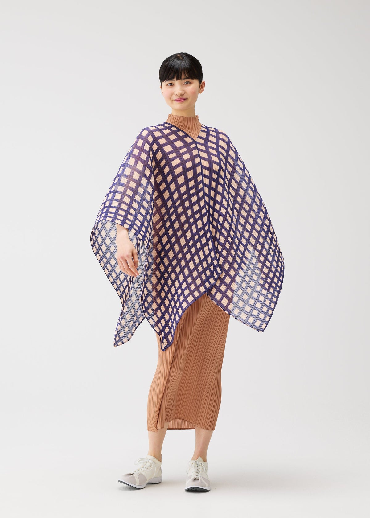 A woman poses against a white background in a tan pleated dress and white sneakers, wearing the PLEATS PLEASE ISSEY MIYAKE BASKET MADAME-T—a purple-and-white checkered, basket-inspired poncho—smiling subtly with one arm raised to show its flowy shape. view 4 of 4