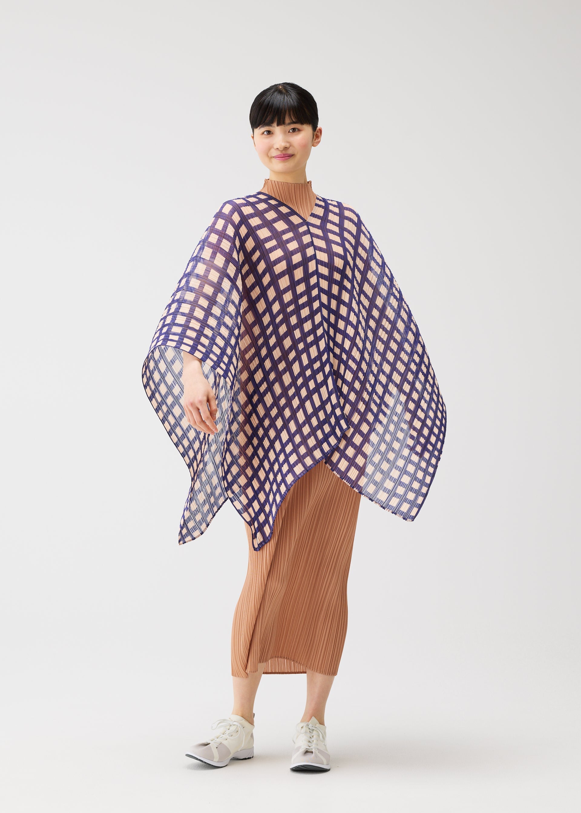 Zoomed view A woman poses against a white background in a tan pleated dress and white sneakers, wearing the PLEATS PLEASE ISSEY MIYAKE BASKET MADAME-T—a purple-and-white checkered, basket-inspired poncho—smiling subtly with one arm raised to show its flowy shape.