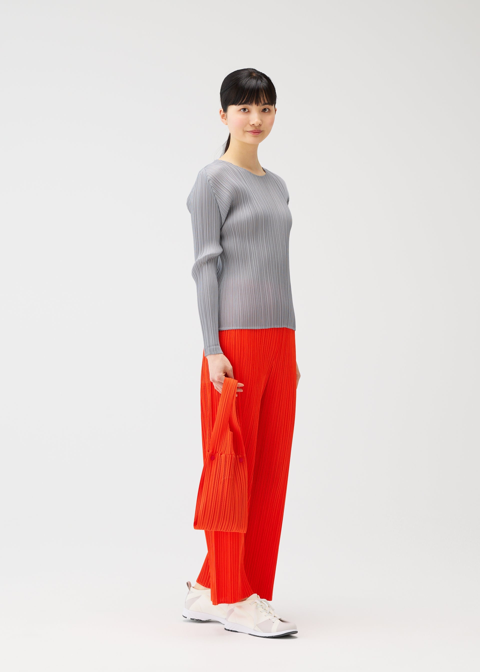 Zoomed view A woman stands against a plain background, wearing a long-sleeve gray ribbed top, bright orange pleated pants, white sneakers, and holding the PLEATS BRIDGE BAG by PLEATS PLEASE ISSEY MIYAKE in her left hand.
