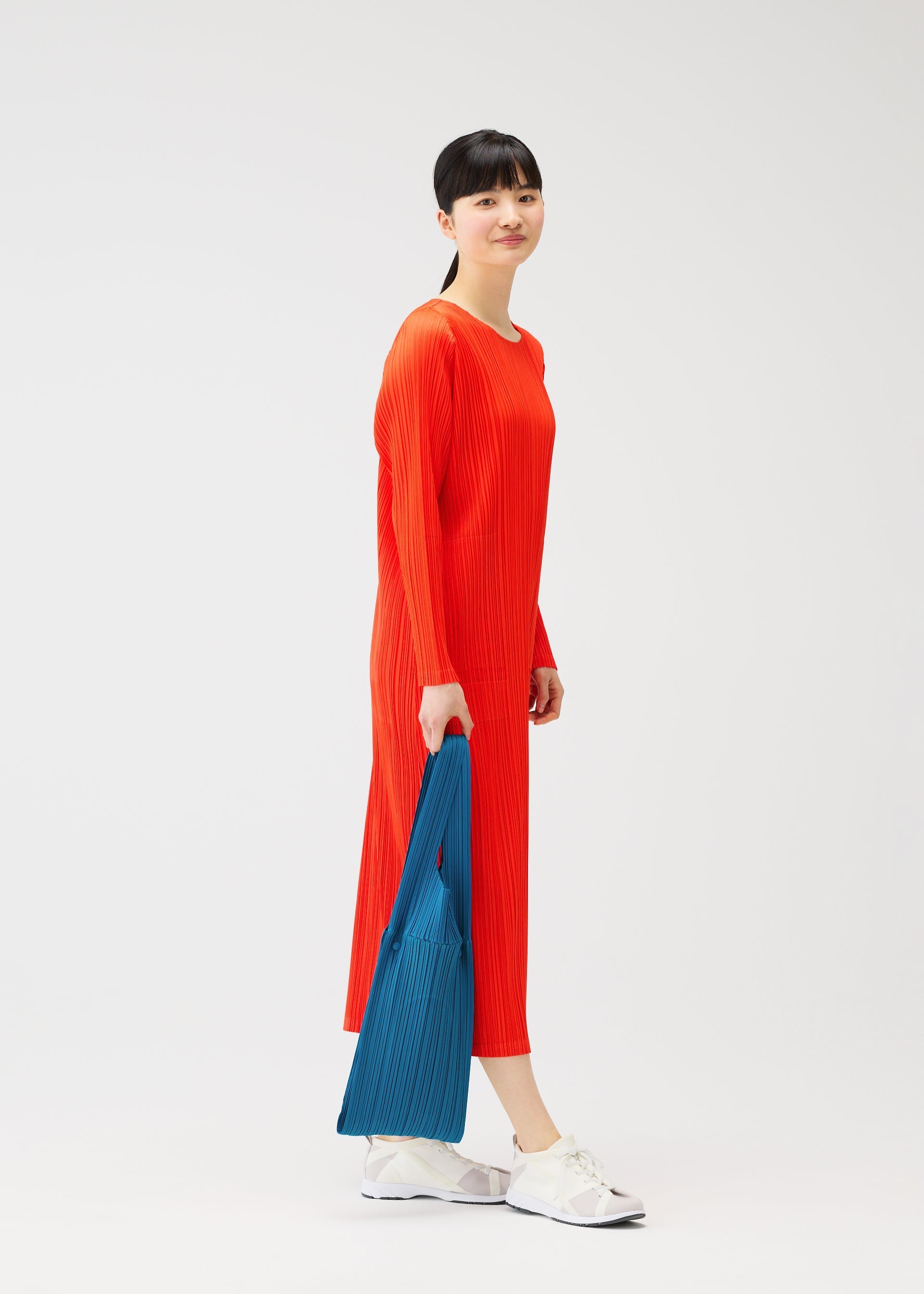 Zoomed view A woman with straight black hair in a ponytail wears a long, bright red dress and white sneakers, holding the PLEATS BRIDGE BAG by PLEATS PLEASE ISSEY MIYAKE. She stands against a plain white background, looking to the side and smiling.